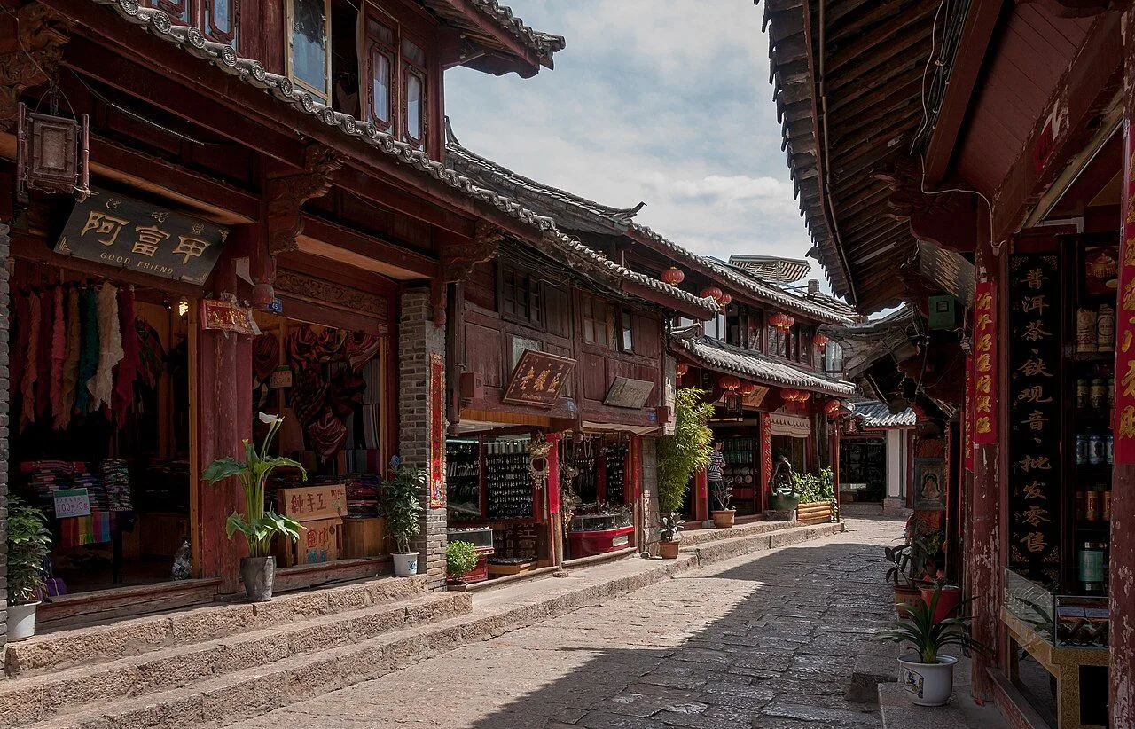 A stone-paved pedestrian street in Lijiang Old Town lined with traditional Naxi heritage shops, the 阿富甲 ("Good Friend") sign visible, red lanterns hanging, and the carved timber facades of the 800-year-old town; the daily-life atmosphere of the UNESCO site