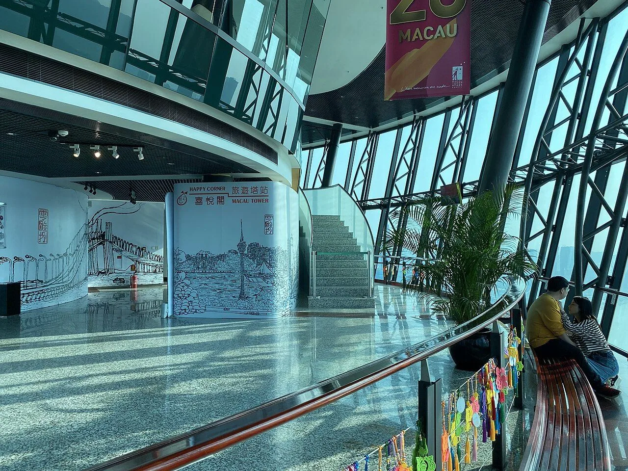 The Macau Tower interior spiral staircase at level 58 with the MACAU 25th anniversary banner hanging from the ceiling, panoramic glass walls revealing the waterfront and the iconic curved silhouette of the tower structure. Built in 2001 by the Sociedade de Turismo e Diversões de Macau