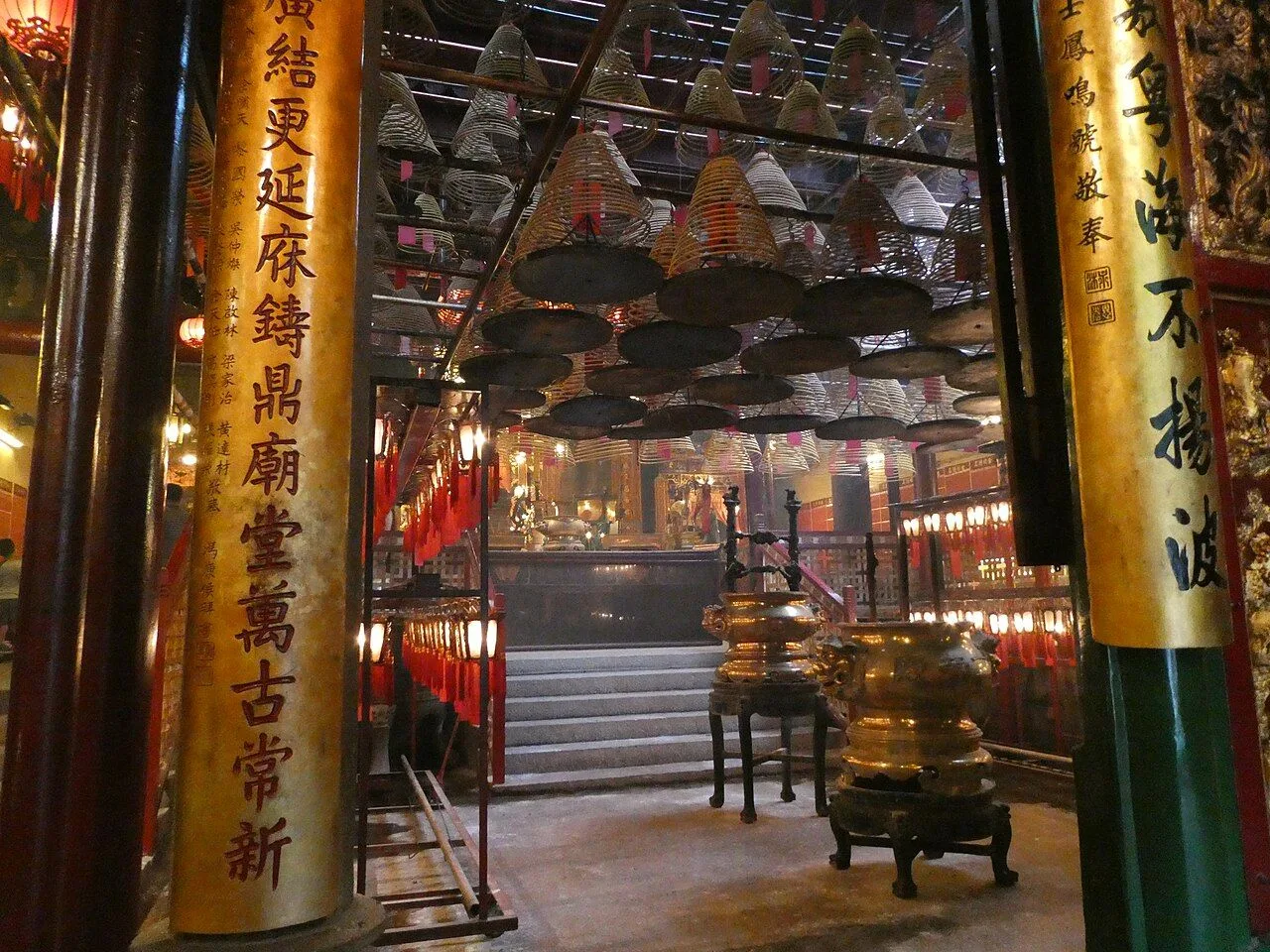 The interior of Man Mo Temple showing the towering incense burner in the foreground, massive yellow pillars with red Chinese calligraphy dedicating the temple to Man Cheong and Mo Tai (the gods of literature and war), and the famous incense coils visible in the background hanging from the dark wooden ceiling