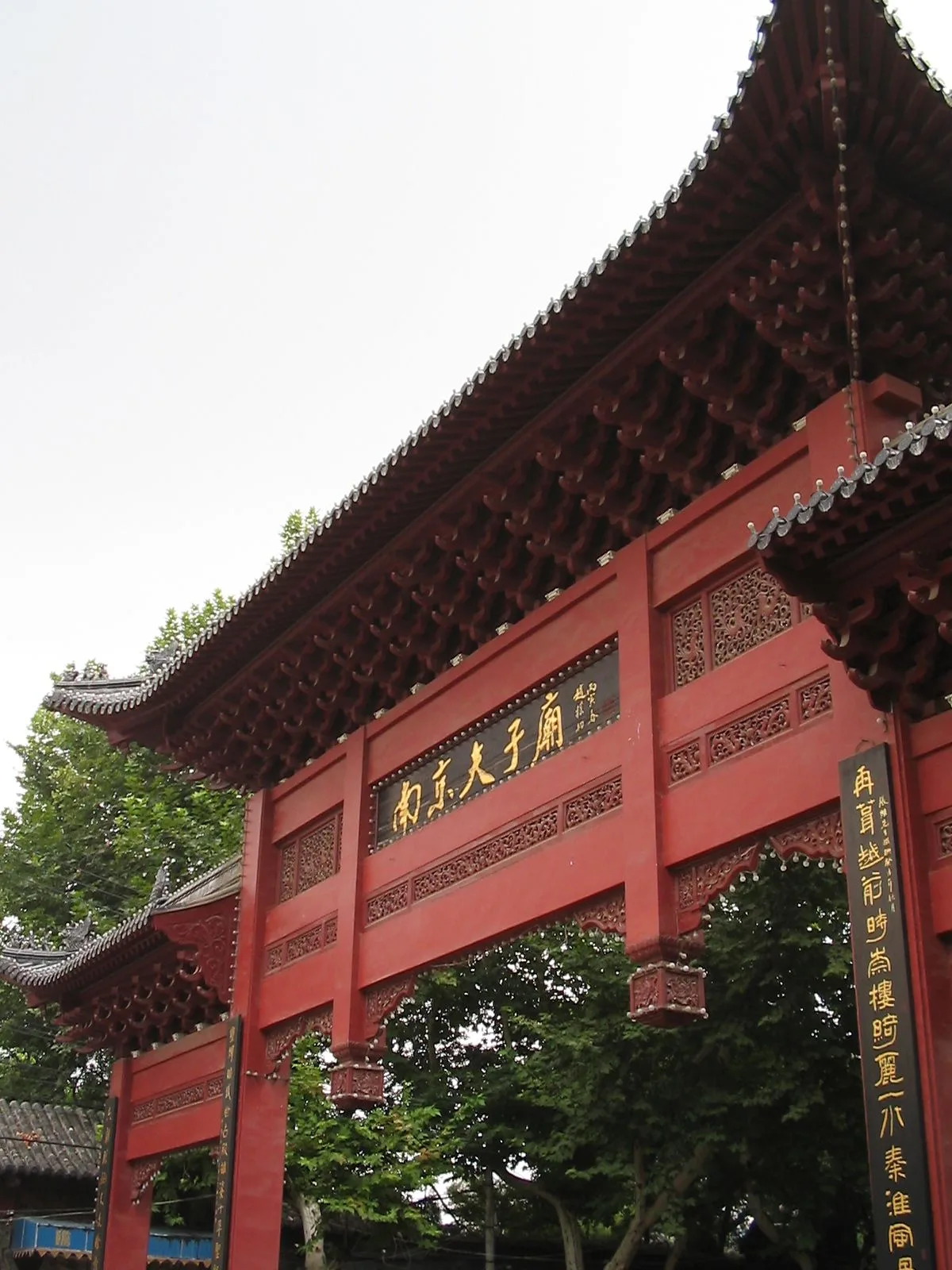 The daytime red wooden gate of the Confucius Temple (Fuzimiao) in Nanjing — the traditional Chinese characters 南京夫子庙 (Nanjing Confucius Temple) inscribed on the gate plaque, the carved wooden beams and red columns of the entrance structure, the green ginkgo trees around the temple, the daytime view that visitors see when they walk into the rebuilt Ming-dynasty Confucian complex from the main street