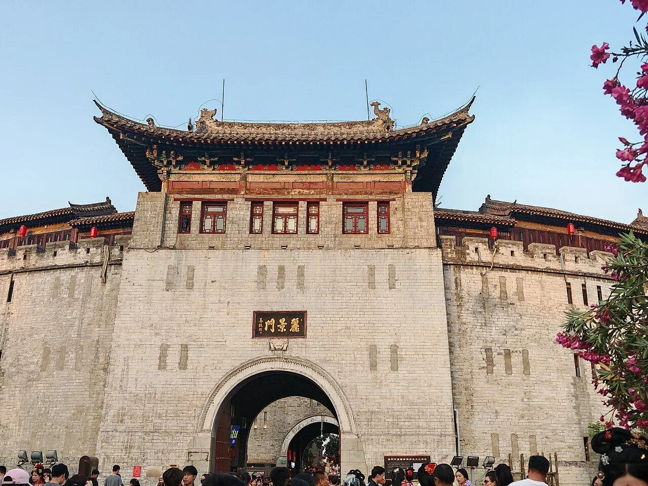 The Lijing Gate of Luoyang in spring 2025 — the ancient three-tiered gate tower with the inner archway visible, surrounded by visitors during the cherry blossom season, the entrance to the food street that anchors the Luoyang Old Town Cross Street (老城十字街) and runs nightly until 11pm