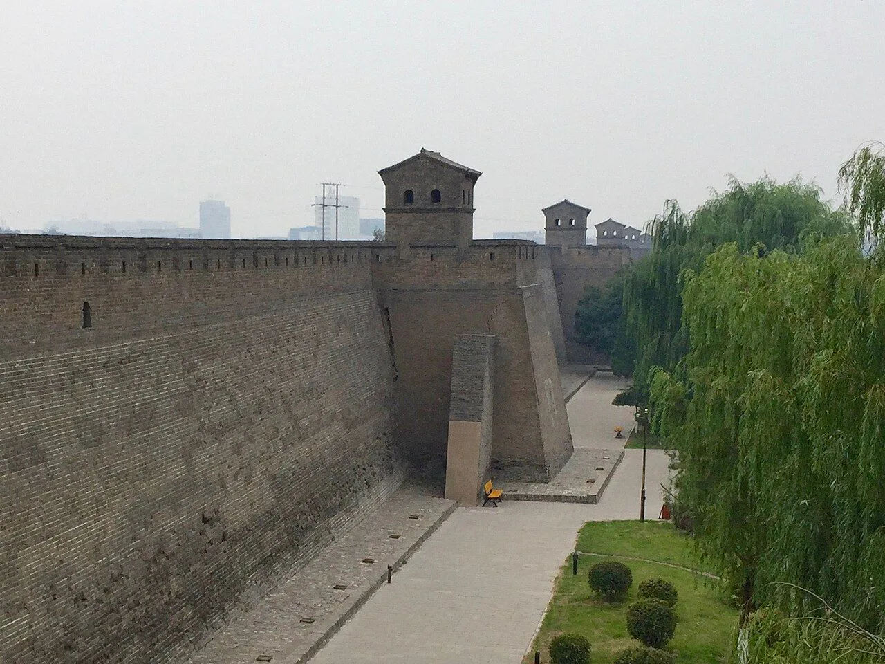 The Pingyao city wall extending into the distance — a continuous line of Ming-dynasty brick battlements punctuated by watchtowers every 50-100m, lined with weeping willow trees on the outer side, the 6km perimeter wall that has encircled this Shanxi merchant city for over 650 years and made Pingyao one of the best-preserved walled cities in the world