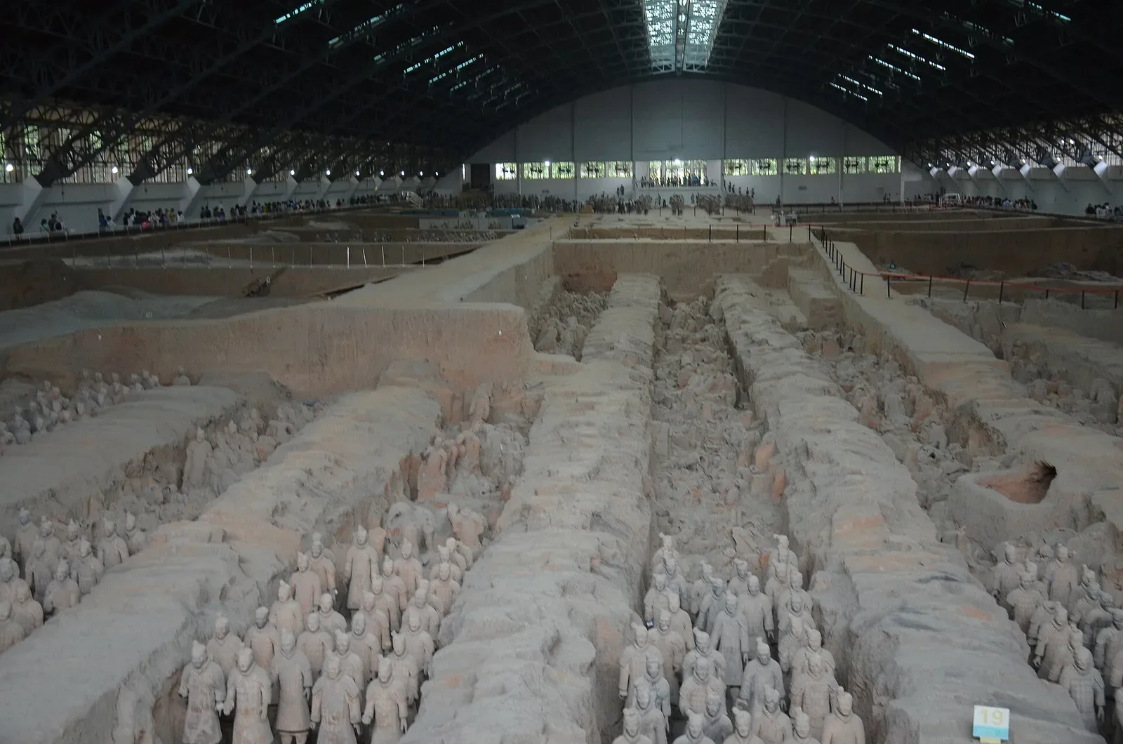 Wide view of Pit 1 from the upper platform showing the full expanse of the excavation hall with the curved roof, the visitor viewing area, and rows of terracotta warriors stretching into the distance under artificial lighting