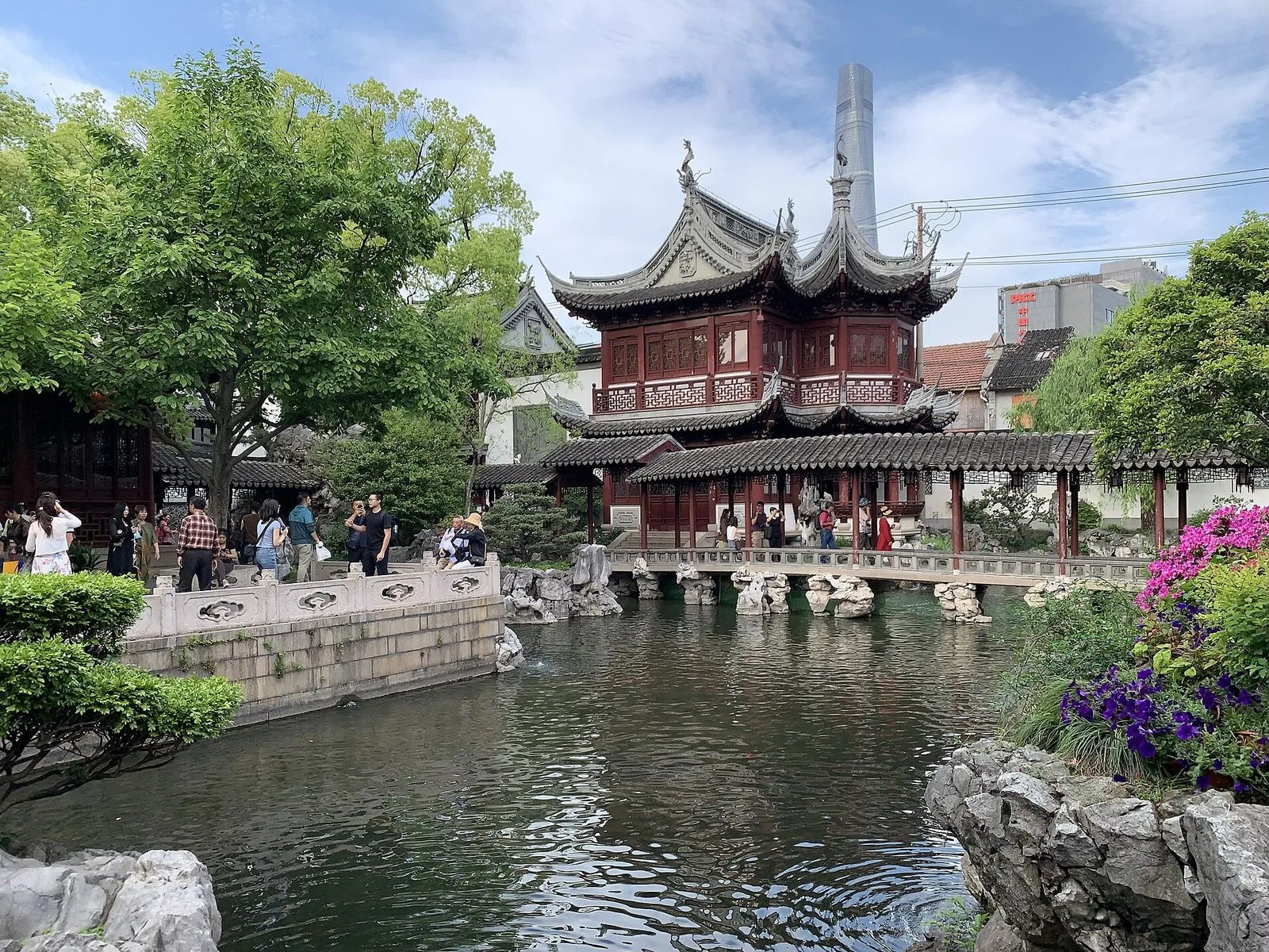 The central pond at Yu Garden with the multi-tiered traditional pavilion in dark red wood and gray-tile roof standing at the far end, visitors walking along the covered walkways on both sides, decorative Taihu scholar rocks placed throughout the garden, and a clear blue sky overhead. A classic example of Jiangnan scholar garden composition with the central water feature as the focal point