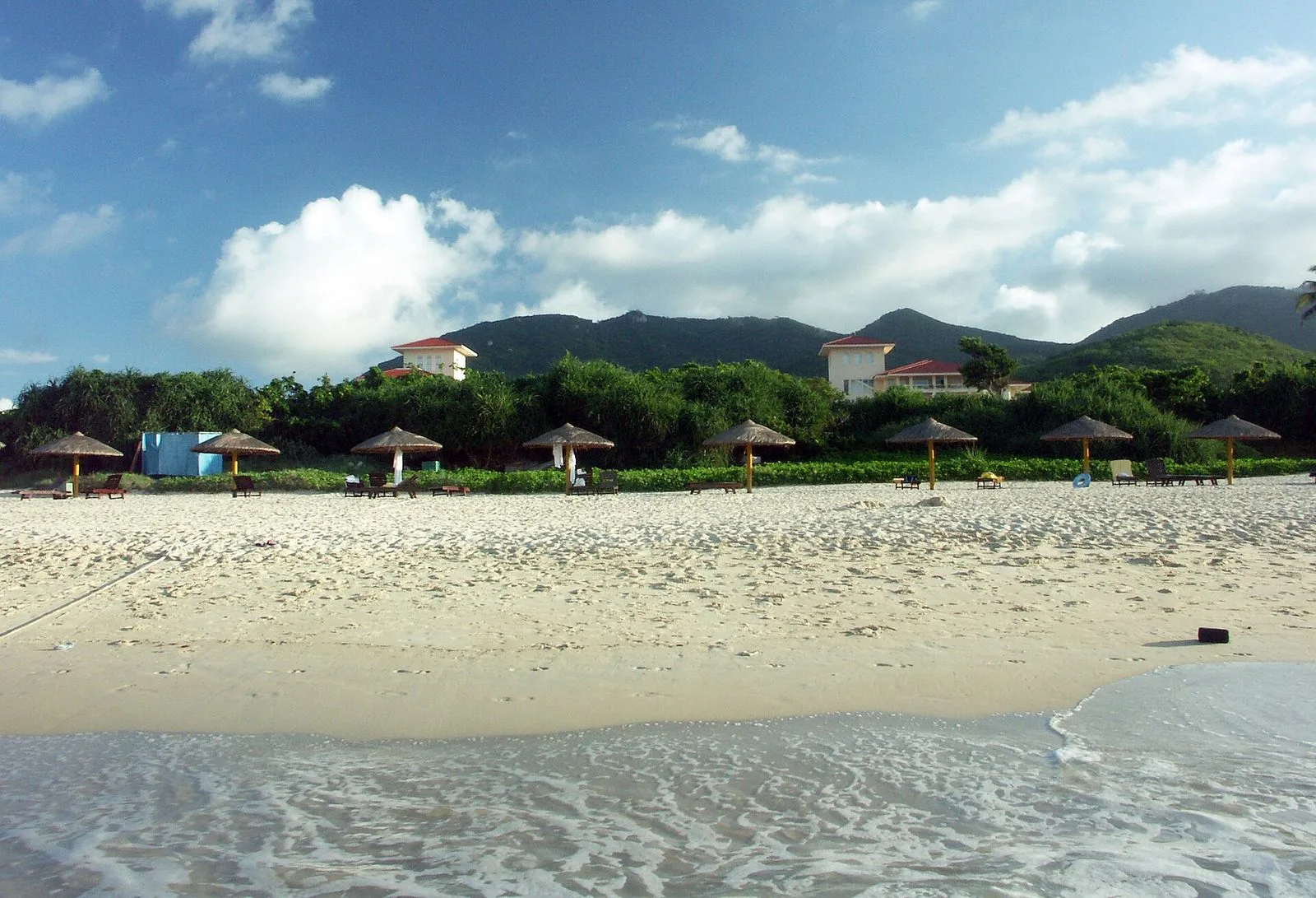 A quiet stretch of Yalong Bay beach lined with thatched sun umbrellas — the empty white sand, blue water, and lush green coastal mountains in the distance, the calm morning scene that makes the bay a popular escape for Hainan residents and visitors from across China who want a tropical beach within a 90-minute flight from anywhere in East Asia