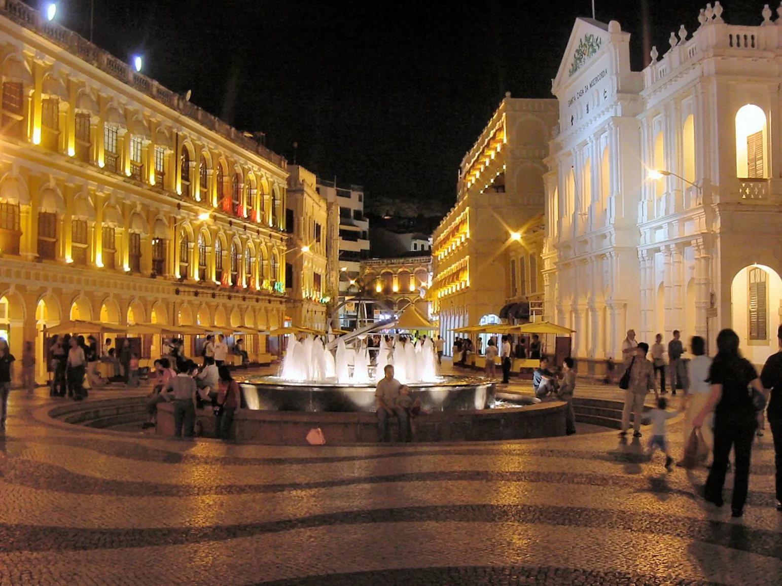 Senado Square at night — the wave-pattern pavement lit from below, the armillary-sphere fountain at the center spraying water, the colonial Portuguese buildings painted in warm yellow and illuminated, the neoclassical white Holy House of Mercy on the right, the night scene that has become the most-photographed view of Macau’s historic heart and the gathering place during the annual Macau International Fireworks Festival
