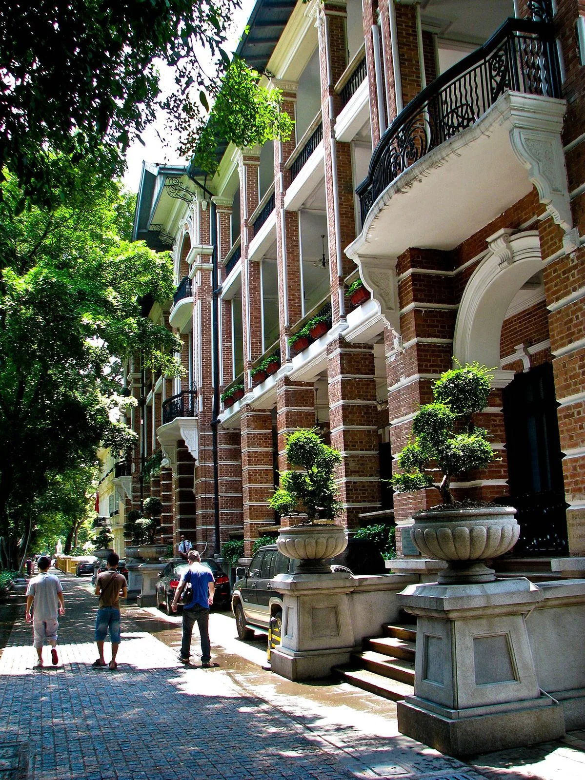 A European-style street on Shamian Island with the 1890s colonial buildings on one side featuring red brick walls, arched doorways, and ornate cast-iron balconies, with three men walking down the cobblestone street away from the camera and cars parked alongside