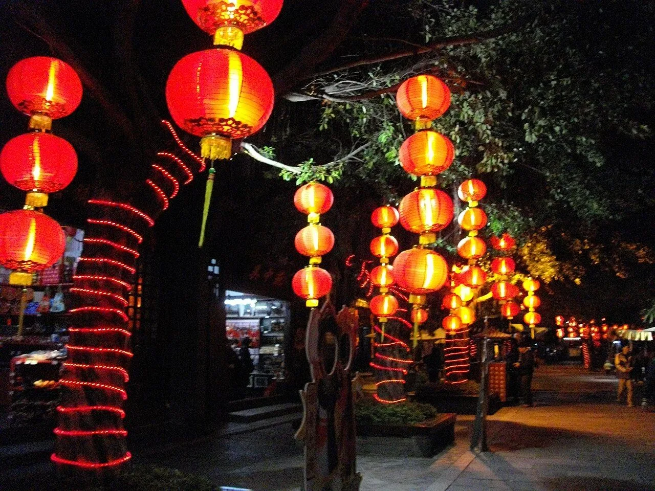 Rows of red Chinese lanterns glowing brightly at night along a pedestrian path in Splendid China Folk Culture Village, the lantern poles wrapped with red LED spirals creating a festive tunnel of light with visitors visible in the distance under the warm glow