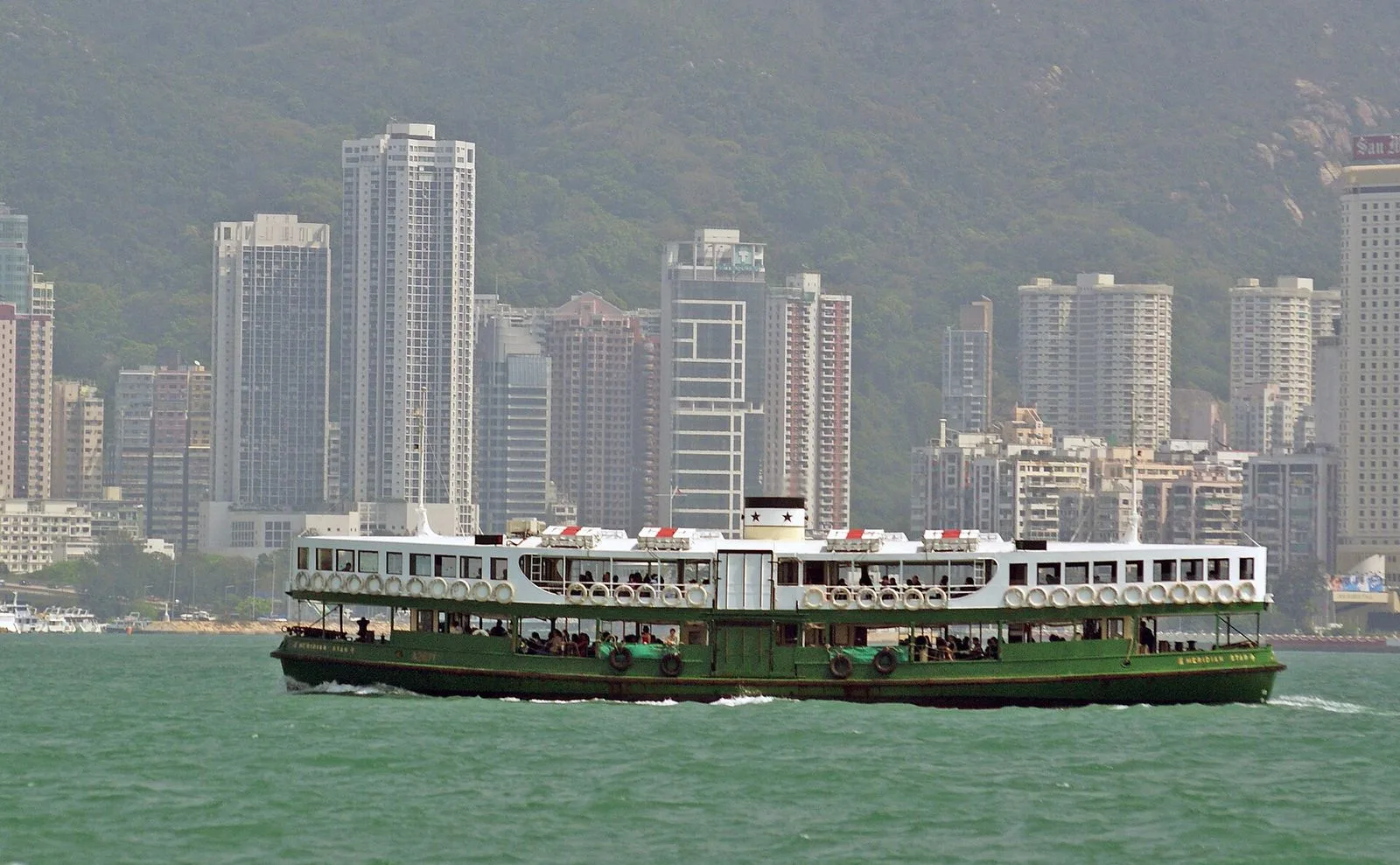 The Celestial Star (天星) ferry crossing Victoria Harbour — the iconic green-and-white double-deck Star Ferry with Chinese characters 天星小轮 and English CELESTIAL STAR painted on the bow, the Kowloon skyline rising behind, the vintage ferry that has plied the harbor since 1888 and is one of the most-photographed boats in Asia, named after the Star Ferry Company founded by Dorabjee Naorojee Mithaiwala in 1888