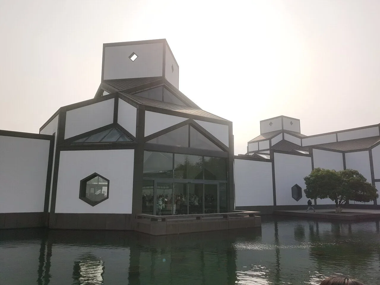 The exterior of the Suzhou Museum seen across the reflecting pool, showing I.M. Pei's signature combination of white walls, dark tile roofs, and hexagonal window openings, with the building's modernist geometry perfectly mirrored in the still water of the central pool
