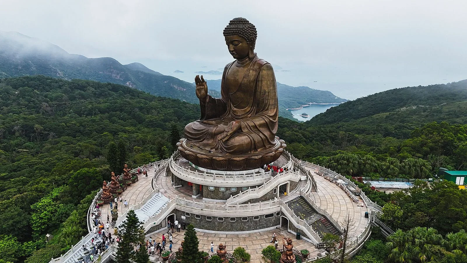 A drone-aerial view of the Tian Tan Buddha (天壇大佛) on Lantau Island — the seated bronze Buddha statue raised on its stone platform above Ngong Ping 360, the circular stone walkway and the 268 steps leading up to the altar, the lush green mountains of Lantau Island stretching into the distance with the South China Sea visible beyond, the Buddhist statue that takes 30,000 visitors a month to climb the 268 steps to reach