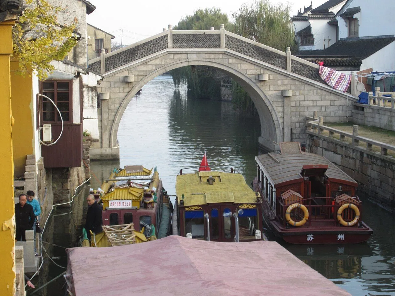 A stone arch bridge over a canal at Tiger Hill, Suzhou, with traditional tour boats passing under the bridge and local residents standing on the left embankment — the classic water-town scene of Suzhou's canal network with whitewashed buildings and willow trees