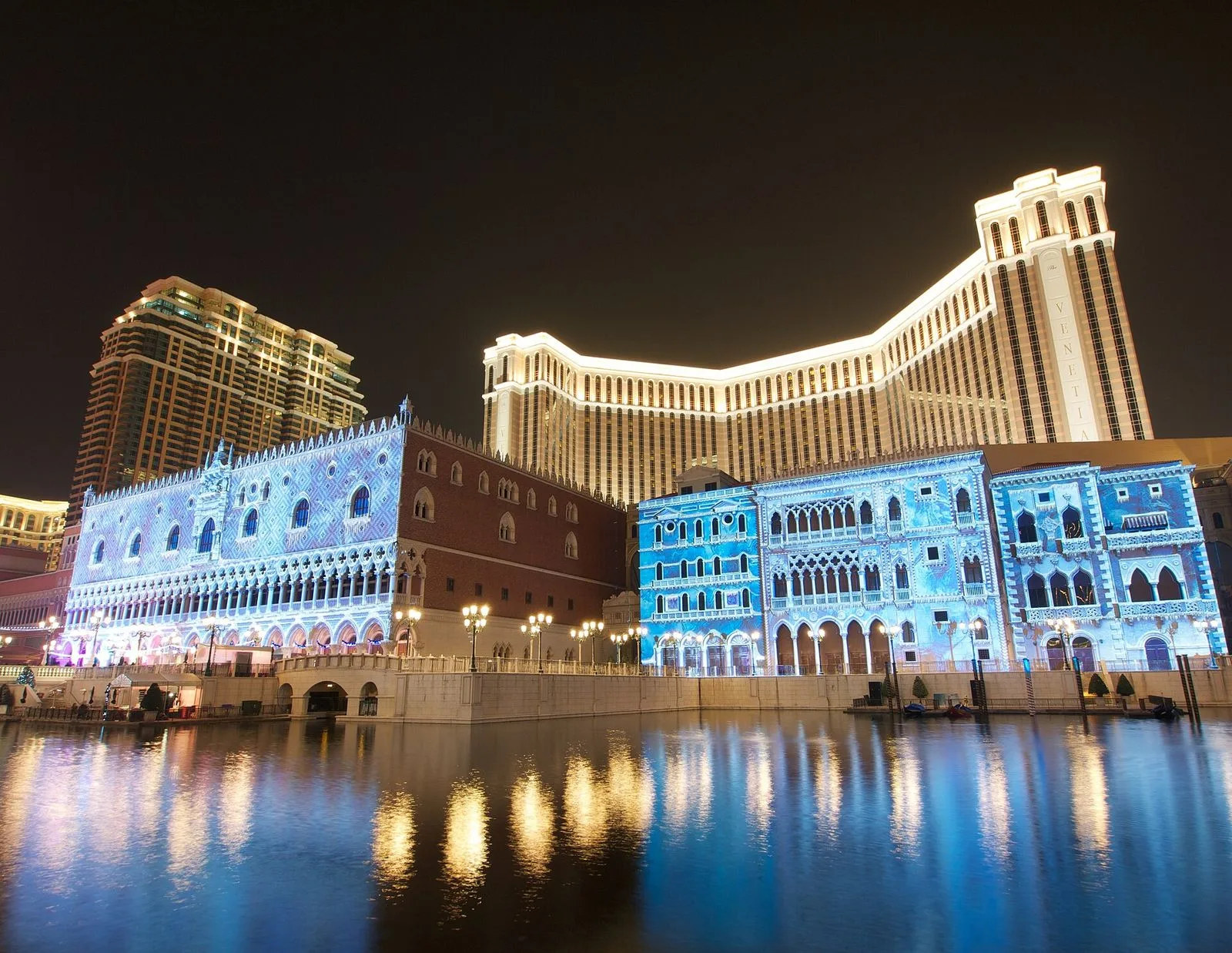 The Grand Canal Shoppes lagoon inside The Venetian Macao at night — the indoor canal with the gondolas, the painted sky ceiling lit in deep blue, the Venetian towers reflected in the water, the evening atmosphere of the resort that operates 24/7, the 3,000-suite hotel tower that rises above the mall, the most-photographed night view inside the resort that anchors the southern end of the Cotai Strip