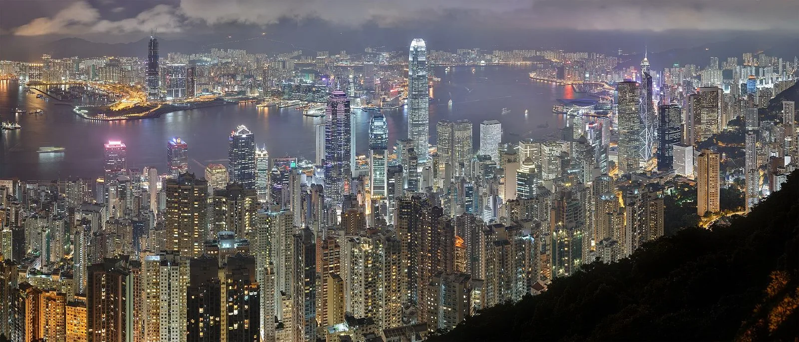 A wider panoramic night view from Victoria Peak — the full sweep of Hong Kong Island skyline from the west (Kowloon visible across the harbour) to the east (Causeway Bay), the thousands of skyscraper lights reflecting in Victoria Harbour, the panoramic view that is best at dusk as the sky turns purple just before the city lights dominate, the 8 million visitors a year who come to the Peak in the evening to watch the city light up