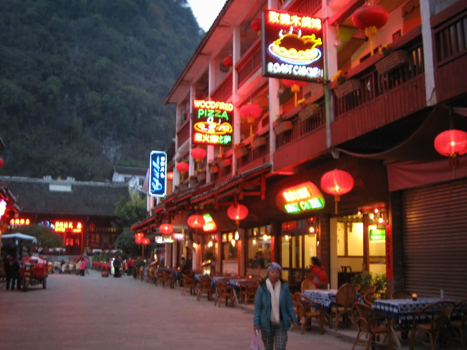 West Street in Yangshuo at dusk — the street lit by red Chinese lanterns and neon signs in both Chinese and English (Wood Fired Pizza, Roast Chicken Inn), the wooden balconies of the restored Ming-dynasty buildings, the cafe tables set out on the stone pavement, the foreigner-hub atmosphere that has been the same for 50 years since the street first opened to Western backpackers in the 1970s, the evening scene that draws Yangshuo's 1.5 million annual visitors for dinner and drinks