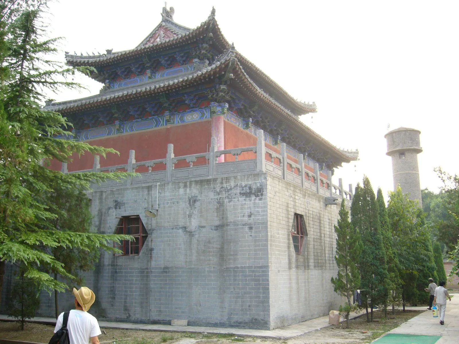 An exterior view of the White Horse Temple showing the gray brick base of a Tang-dynasty hall with the traditional Chinese roof above and the brick Sutra Pagoda (齐云塔, Qiyun Pagoda) visible in the background to the right — the 13-story brick pagoda that has stood in the temple grounds since the Song dynasty (1175 AD), the early Chinese Buddhist architecture that makes White Horse Temple a direct line back to the introduction of Buddhism to China in the 1st century AD