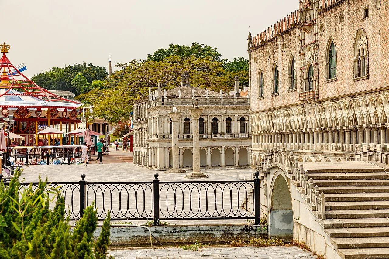 The Italian piazza at Window of the World Shenzhen with a 1:3 scale replica of the Doge's Palace in Venice, the detailed white marble arches and columns, a small carousel with a red top, and a few visitors walking across the open stone piazza under trees