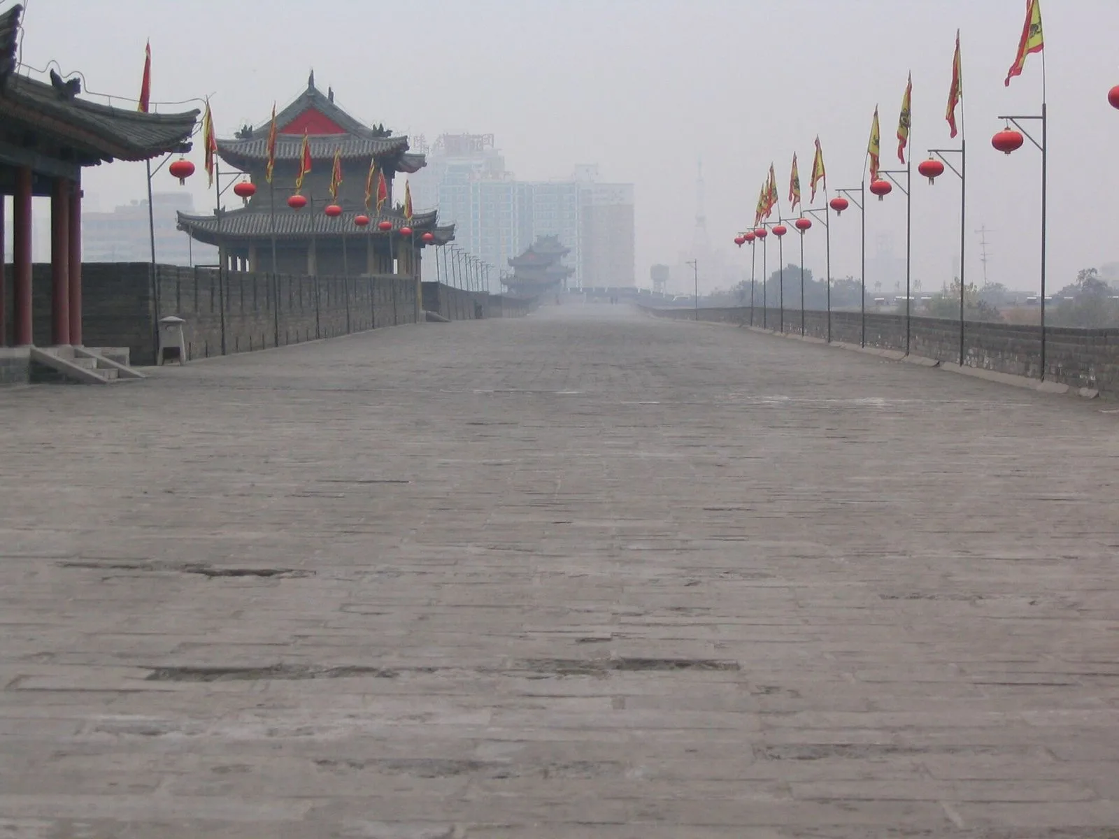 A wide-angle view from on top of the Xi’an city wall — the broad stone-paved battlement wide enough to drive a carriage on, the watchtower at the end of the wall section, the red lanterns and yellow flags lining the parapet, the view that visitors experience when they rent a bike and cycle the full 14 km around the ancient city, the experience that takes 2-3 hours and shows the old city from the unique Ming-dynasty perspective