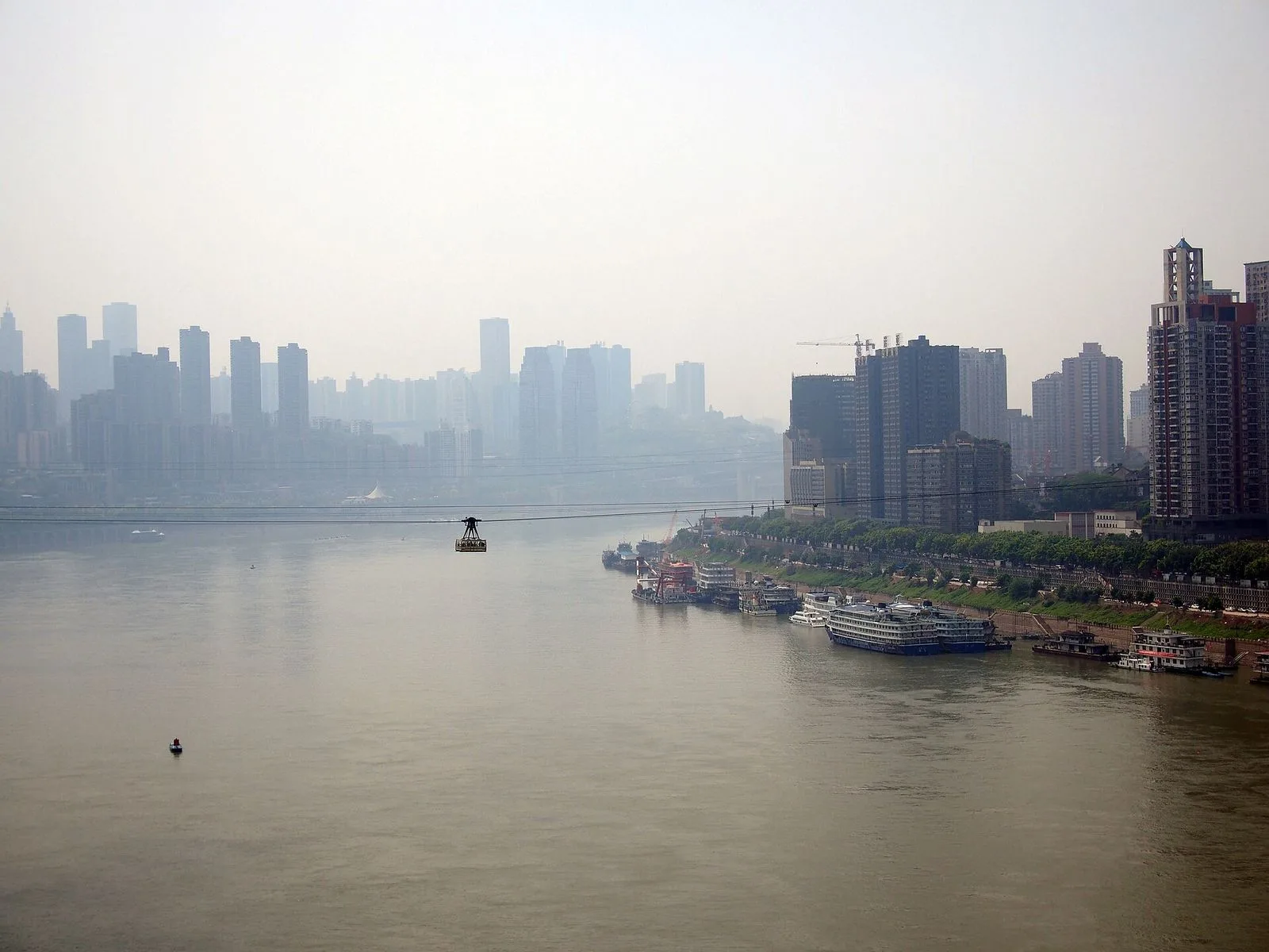 The Chongqing Yangtze River Cableway cabin crossing the river with the cityscape behind — the small cable car suspended from the cables against the hazy view of the city, the cargo ships and riverboats on the Yangtze below, the dramatic backdrop of Chongqing’s iconic skyline rising behind, the cable car that has crossed this stretch of the Yangtze since 1987 and now operates as both commuter transport and tourist attraction