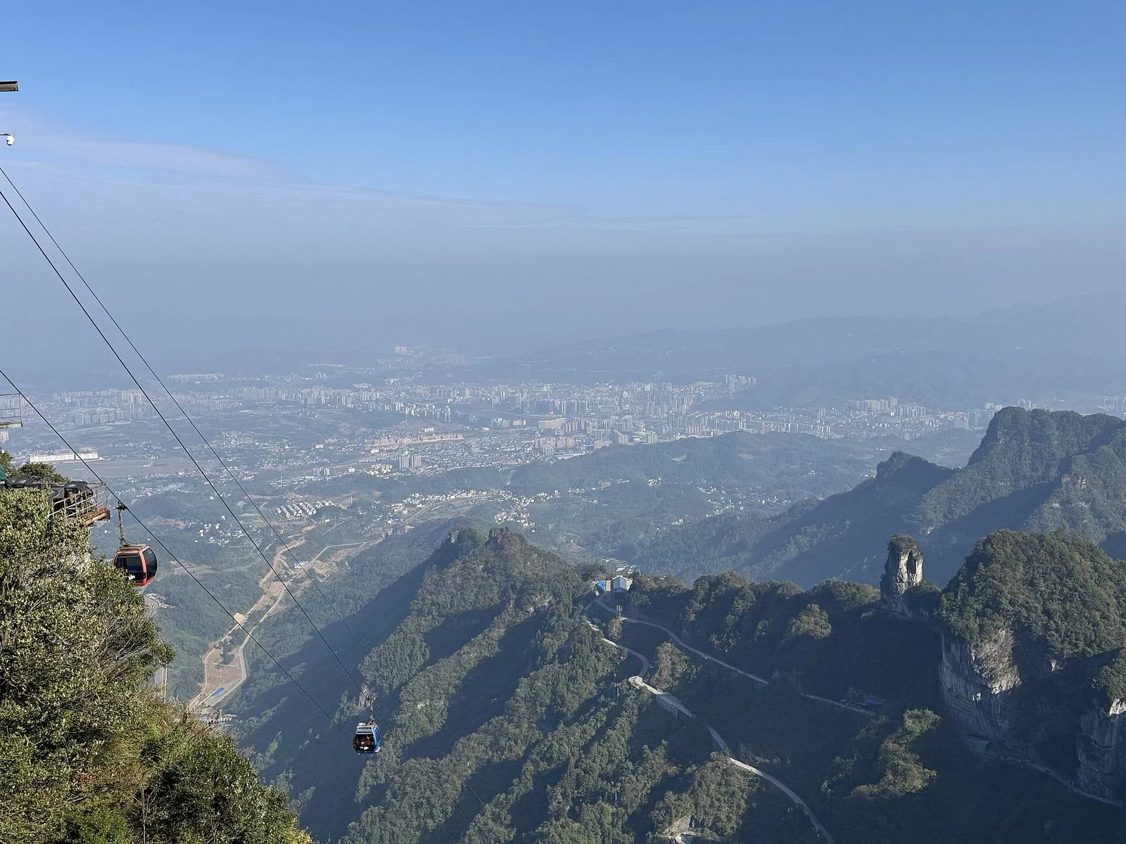 A view from the Tianmen Mountain cable car showing the world’s longest passenger cableway descending into the valley — the red and blue cable car cabins suspended from the steel cables with the city of Zhangjiajie and the surrounding mountains in the distance, the 7,455m cableway that runs from the city center up to the top of Tianmen Mountain, the engineering marvel that takes 28 minutes to climb the 1,279m vertical rise