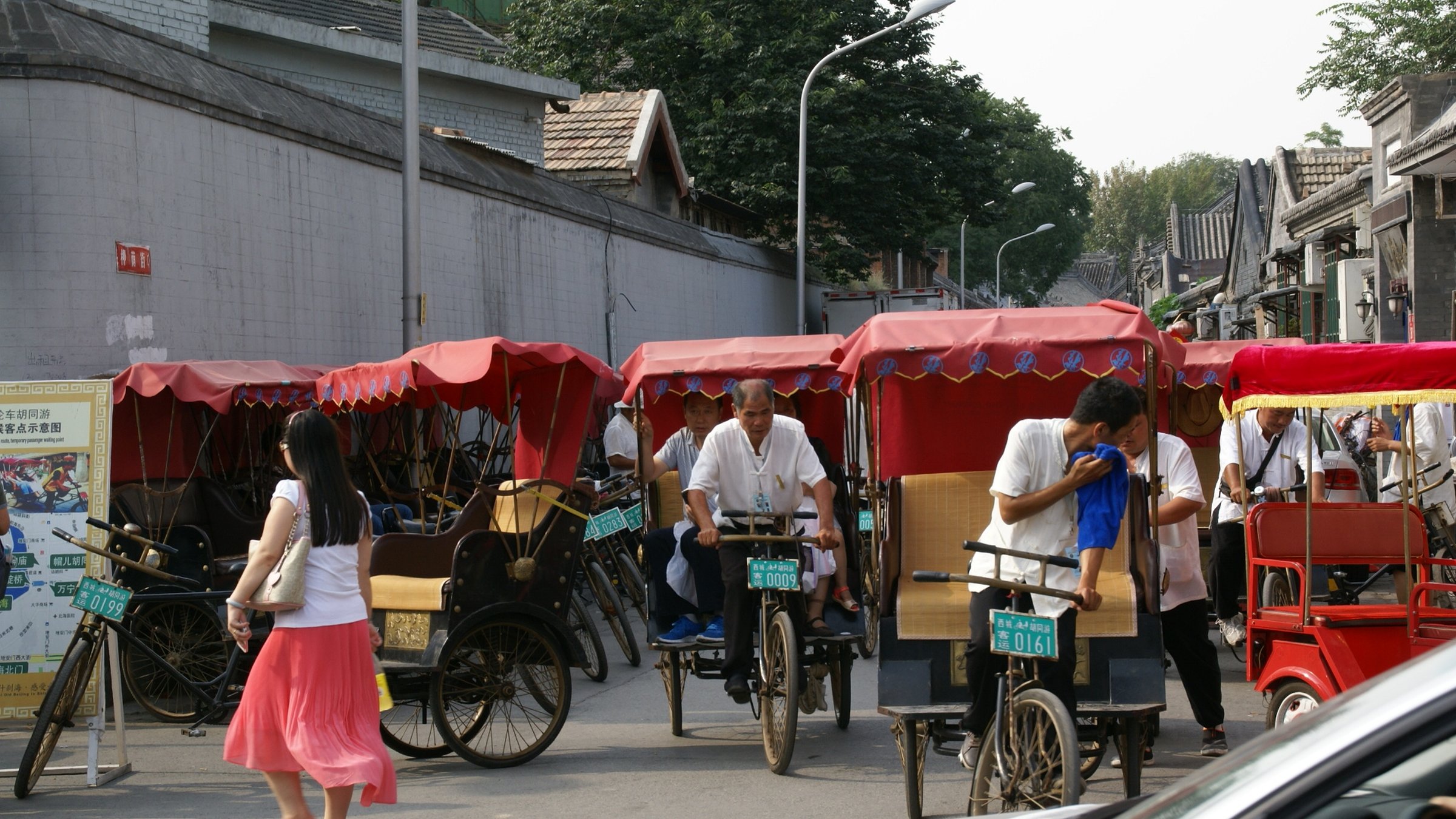 Row of traditional red-canopied Beijing rickshaws (pedicabs) waiting for tourists on a hutong street, with drivers in white shirts and a woman in a pink skirt