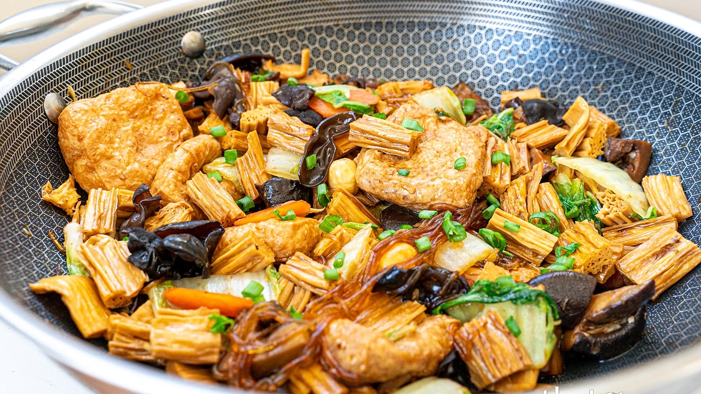 Eine chinesische vegane Pfanne aus Tofu, Waldohrpilzen, Glasnudeln, Pak Choi und Yuba (Tofu-Haut) im Wok – ein typisch buddhistisches Gericht ohne Fleisch oder Eier, glitzernd mit Soße und garniert mit Frühlingszwiebeln
