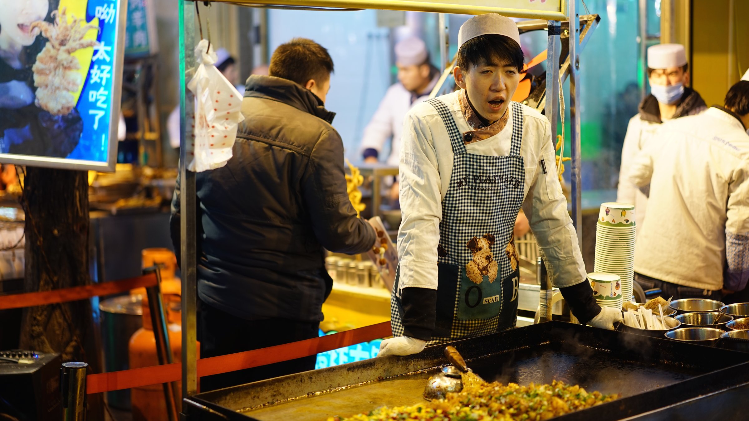 China food by city — a young Chinese chef cooking street food at a night market stall in Xi'an, with a crowd of locals in the background