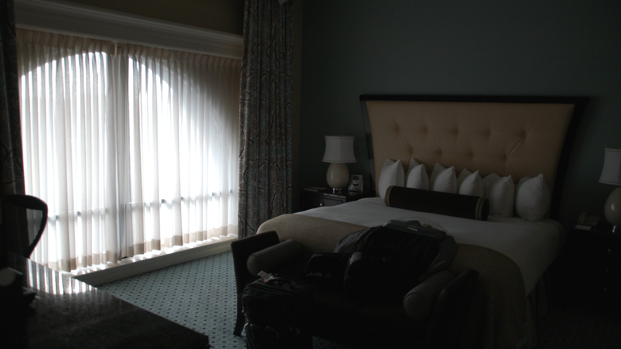 A luxurious modern hotel suite bedroom with a king bed, large window, and elegant furnishings in soft natural light