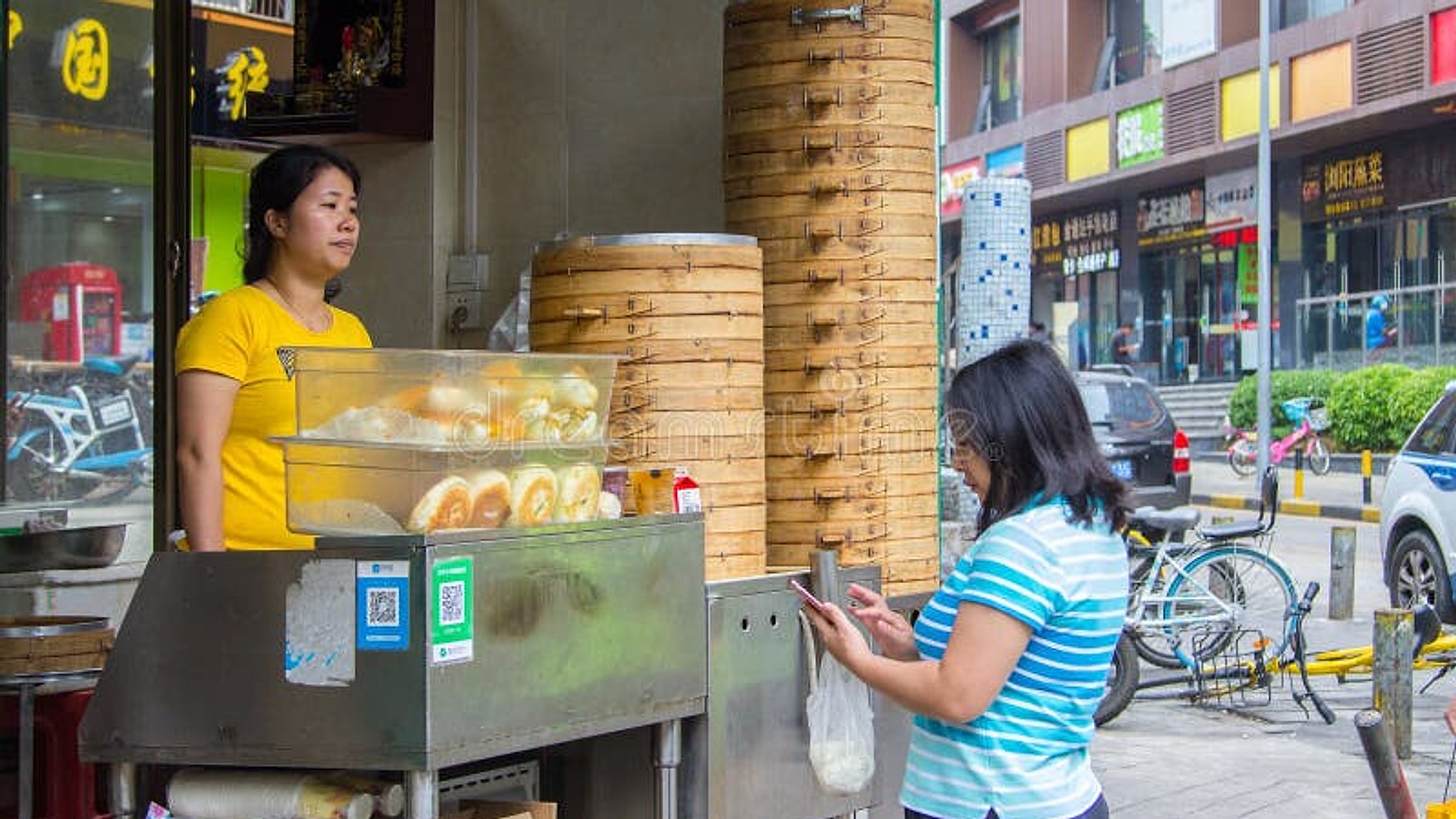 Véritable stand de baozi chinois avec le vendeur montrant deux codes QR WeChat Pay et Alipay laminés sur des cartes imprimées, prêts à être scannés par un client ; cuiseurs à vapeur en bambou empilés visibles derrière le comptoir