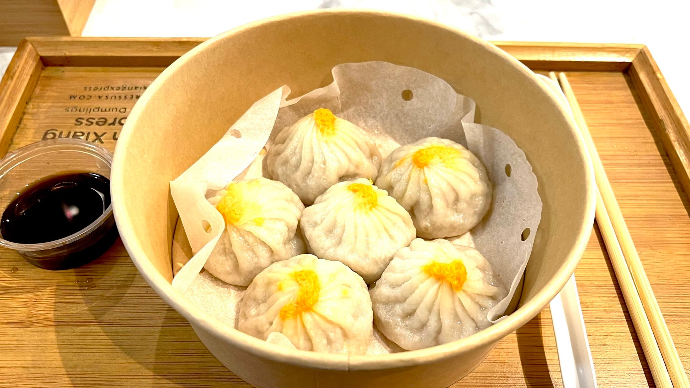 Bamboo steamer of Shanghai-style xiaolongbao soup dumplings with a small dish of black vinegar dipping sauce on a wooden tray — six plump dumplings with the characteristic pinched top, glistening with broth inside the thin translucent wrapper