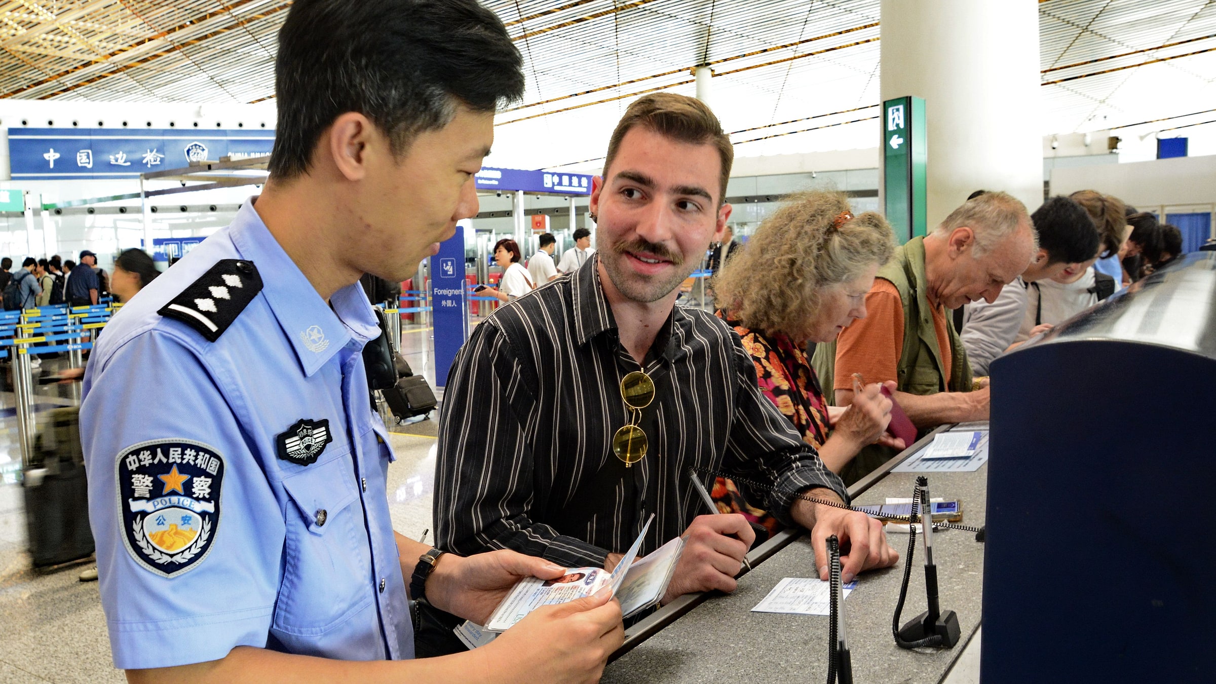 Chinese police officer in the light-blue Public Security Bureau (中华人民共和国警察) uniform helping a Western traveler with documents at an airport/border-control desk — the officer has visible rank insignia and a POLICE patch on the sleeve; the scene captures the approachable, helpful nature of Chinese tourist police and the everyday reality that most foreign-Chinese police interactions are friendly, not adversarial