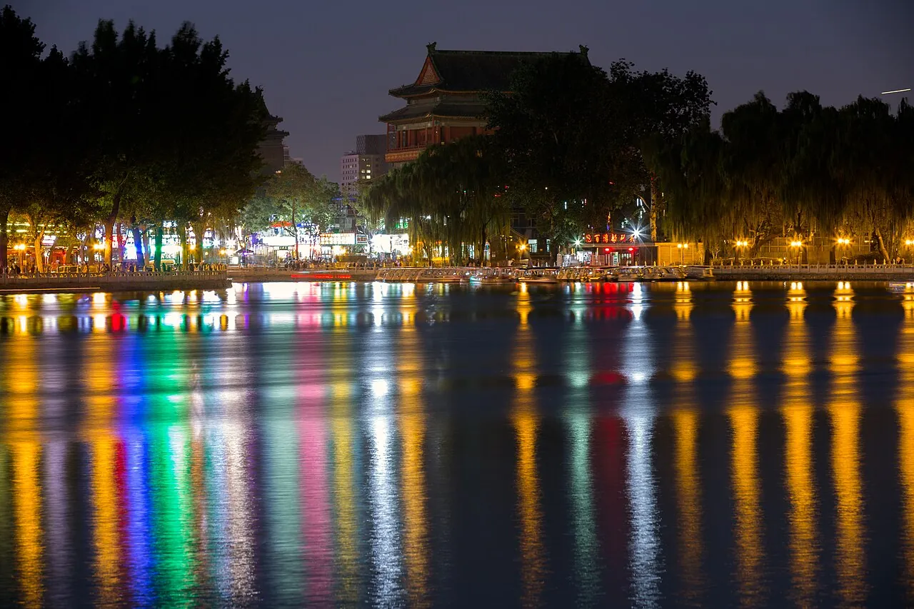 Houhai Lake in central Beijing at night — the calm water reflecting the colorful lights of the surrounding bar-and-restaurant district, the silhouette of willow trees along the lakeshore, the Drum Tower and Bell Tower visible in the distance and a few pedestrians on the lakeside walkway, the most popular nightlife area in the old city