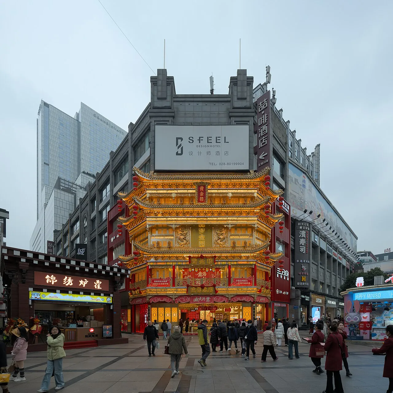 A gold-and-red multi-tiered traditional-style tower glowing on Chunxi Road, with shoppers strolling the pedestrian plaza in front and a SFEEL Designer Hotel sign overhead.