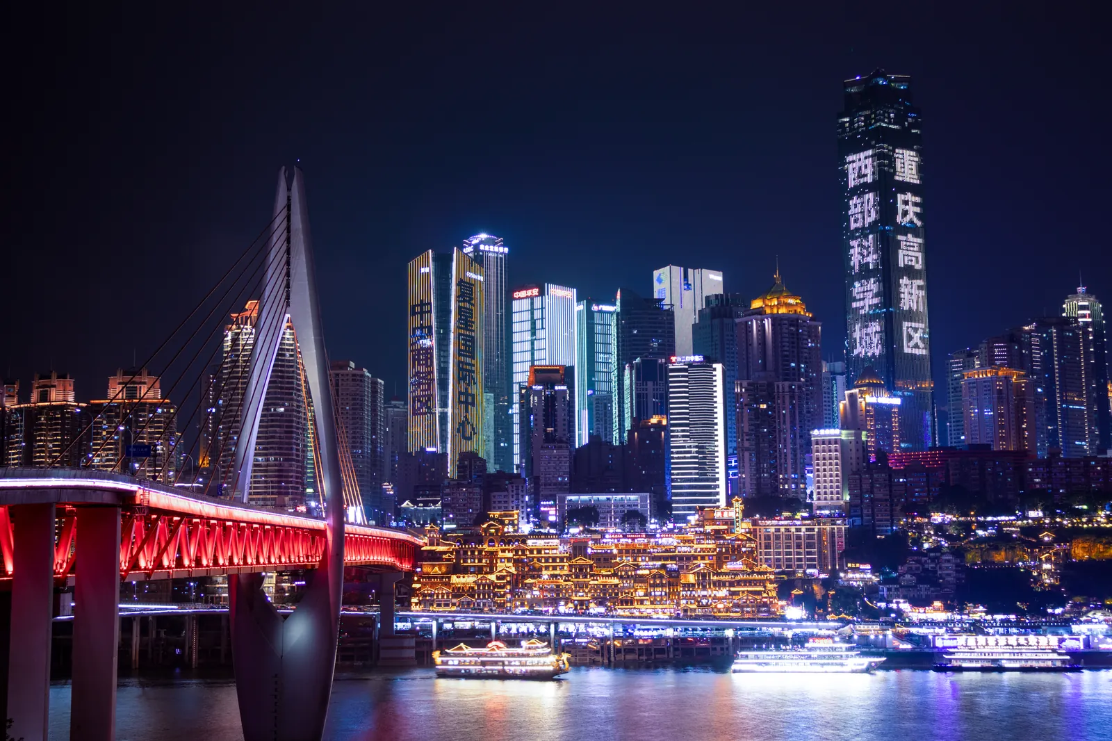 The Chongqing night skyline over the Yangtze and Jialing rivers, with the glowing Hongya Cave complex on the riverbank and a forest of skyscrapers lit up behind, including the red illuminated Qiansimen Bridge on the left.
