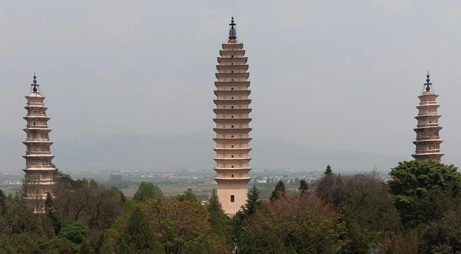 The Three Pagodas of Chongsheng Temple in Dali, Yunnan — three tall white pagodas of different heights standing in a line on a green hilltop, with the Cangshan Mountain range in the hazy distance and the Erhai Lake basin and town of Dali spread out below under a soft grey sky.