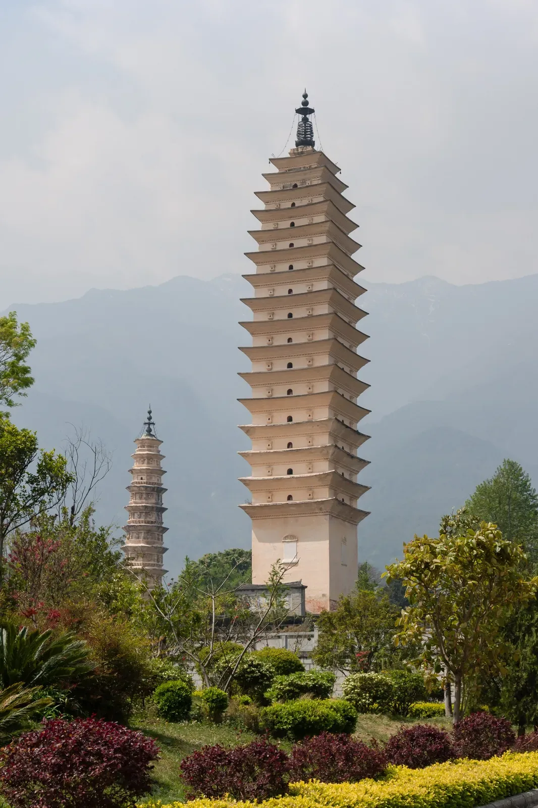 A close-up of the main (tallest) pagoda of the Three Pagodas of Chongsheng Temple in Dali, Yunnan — a 16-tiered white brick pagoda with upturned eaves, a brass finial at the top, and a small shrine at its base, set against a green garden with red-leaf shrubs, flowering trees, and the Cangshan Mountain range in the background.