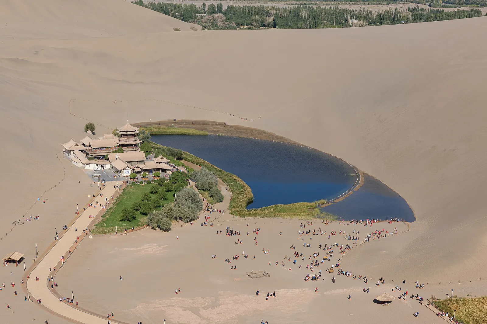 An aerial view of Crescent Lake (Yueyaquan) near Dunhuang, an oasis pond in the shape of a crescent moon nestled among towering sand dunes, with a traditional Chinese pavilion and a cluster of wooden buildings on the shore, and dozens of visitors walking on the sand.