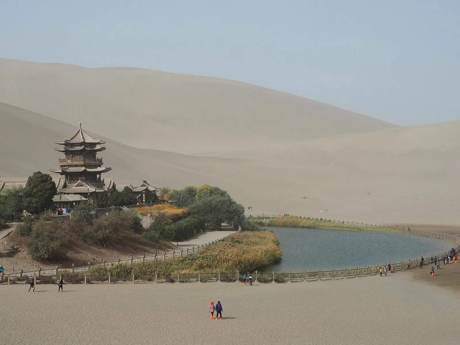 The Crescent Lake (Yueyaquan) oasis near Dunhuang, with its small blue pond framed by golden sand dunes, a multi-storey Chinese pavilion on the shore, and a few visitors on the wooden boardwalk.