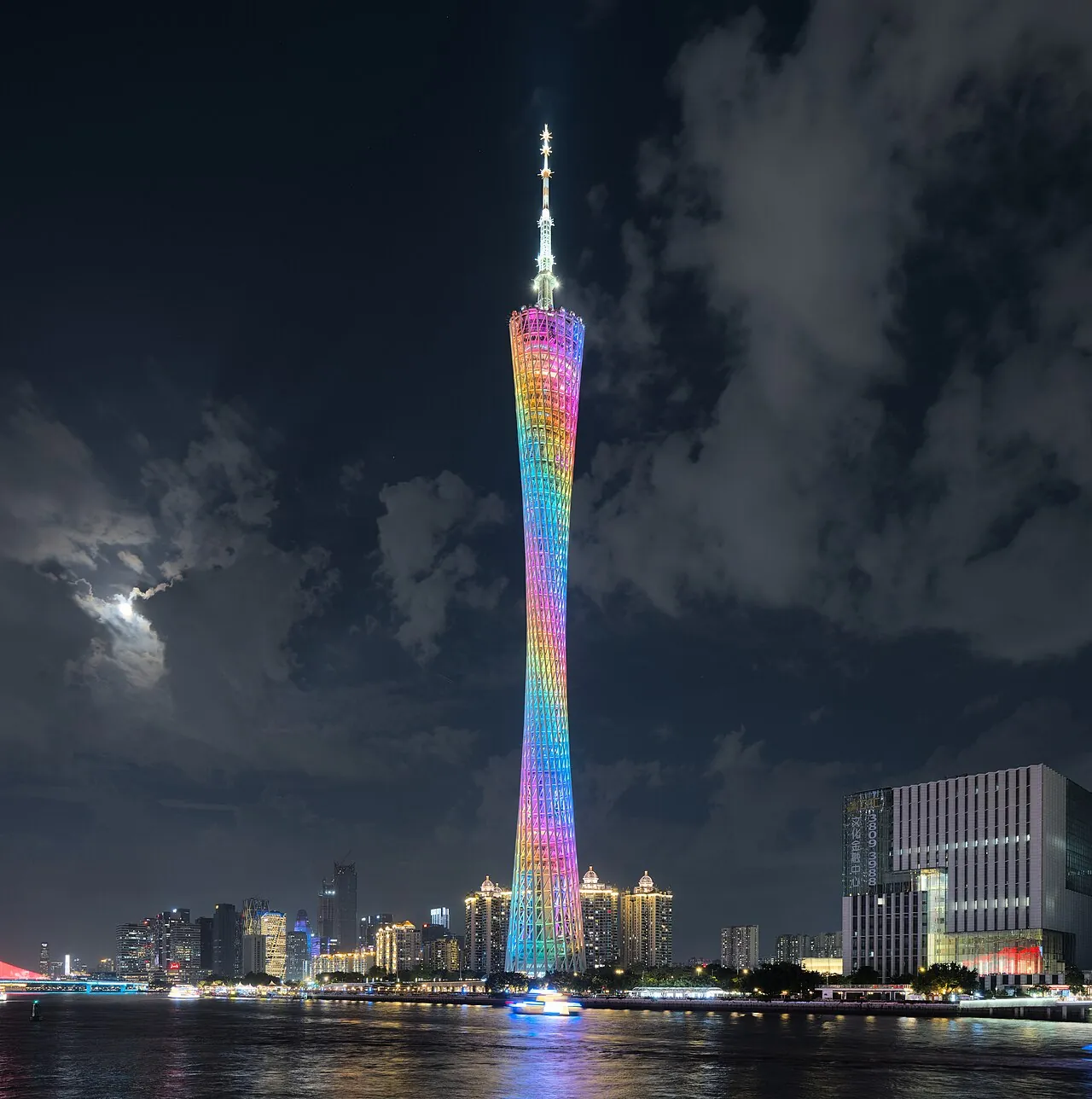 The Canton Tower lit up in rainbow LED at night, with a tourist sightseeing boat cruising the Pearl River in the foreground and a full moon behind clouds.