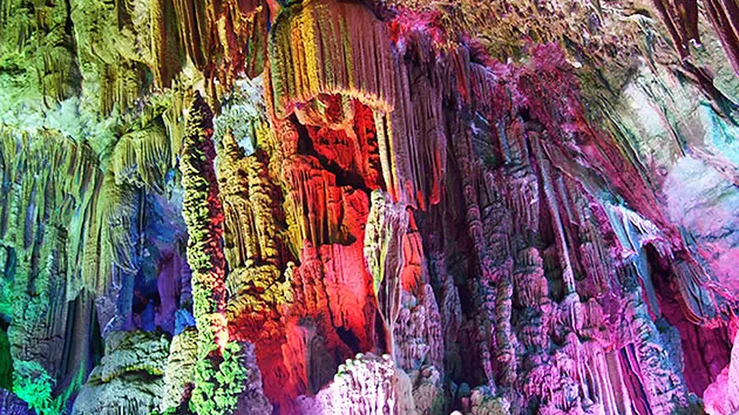 Reed Flute Cave interior — illuminated stalactite columns in red, blue, and green LED lights, with reflection in the cave's underground pool