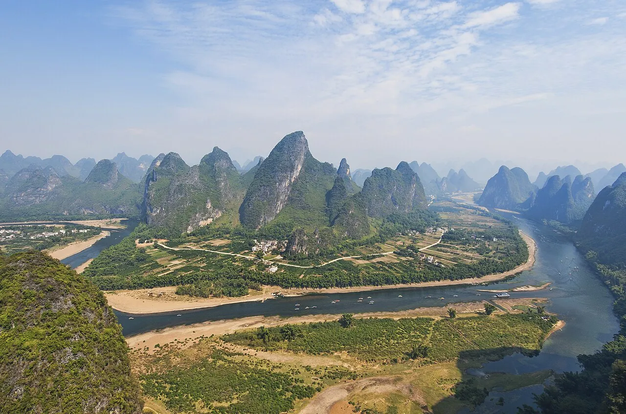 An aerial panorama of the Li River near Guilin, showing a sharp bend in the jade-green water with traditional bamboo rafts, surrounded by forest-covered karst peaks stretching to the horizon under a hazy blue sky.