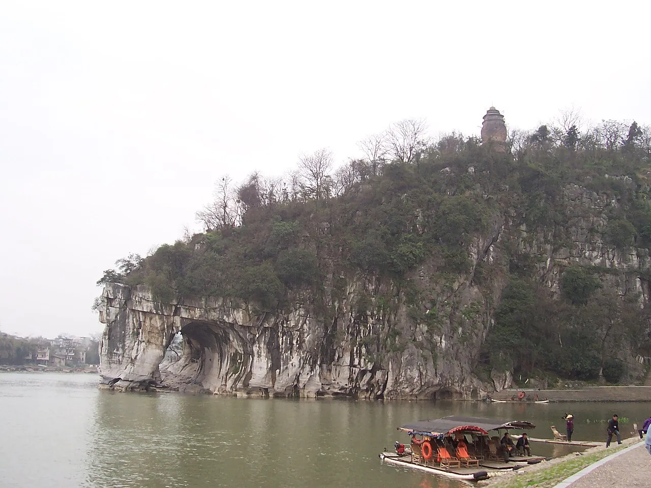 Elephant Trunk Hill in Guilin on a misty day, the karst formation resembling an elephant drinking from the Li River, with two traditional bamboo rafts moored in front of it and a pagoda on the hilltop.