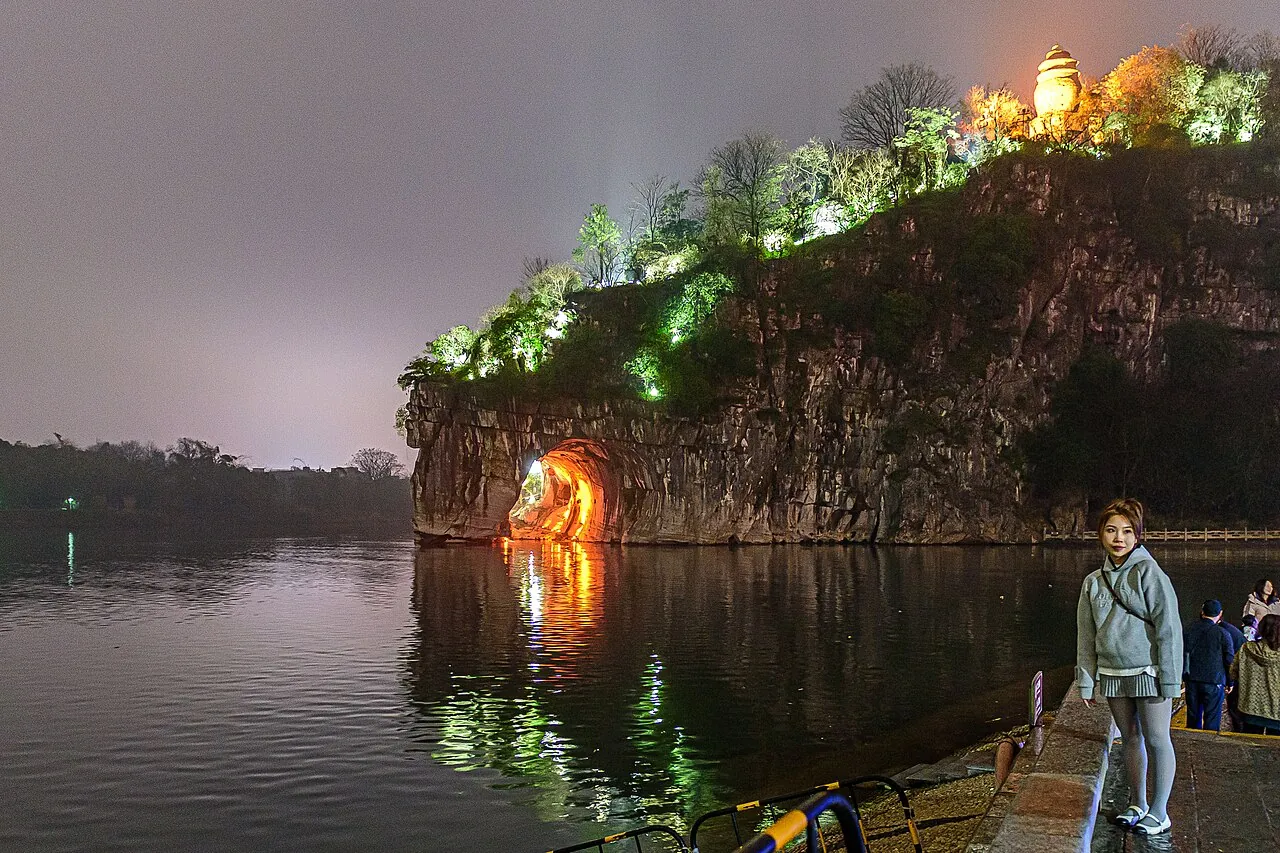 Elephant Trunk Hill in Guilin at night, the archway through the elephant's body glowing with warm orange light and reflecting on the still river, with a local woman standing on the riverbank in the foreground.