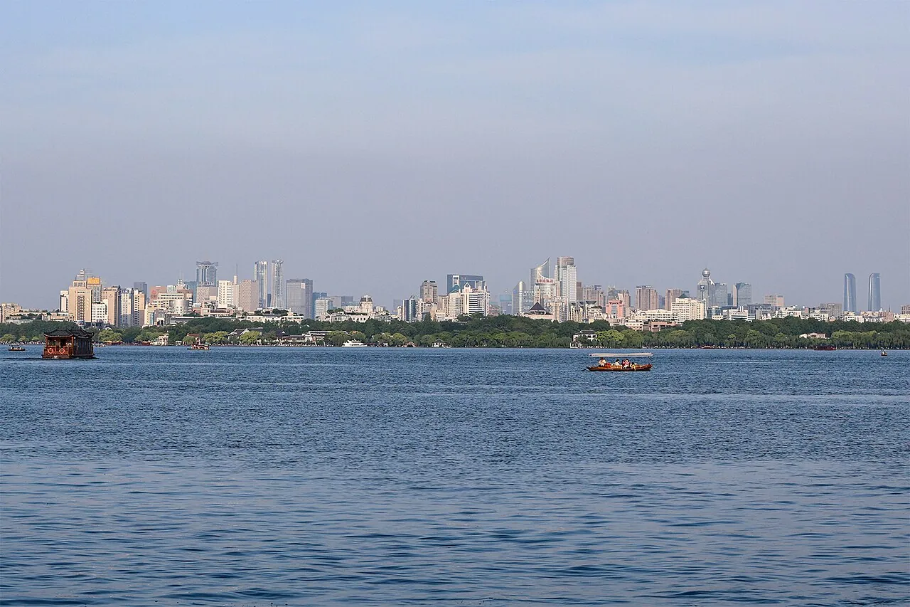 West Lake in Hangzhou on a clear day, with tourist rowing boats on the calm water and the city skyline rising behind a green lakeside park.