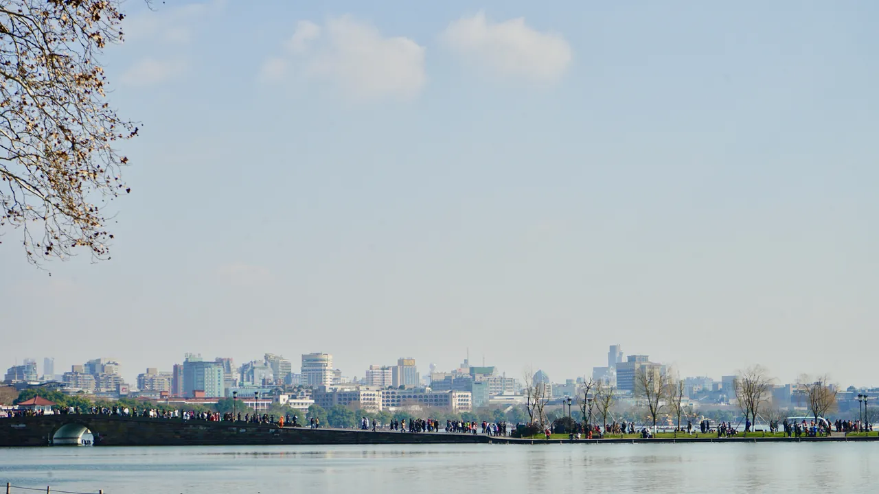 The Su Causeway embankment on West Lake in Hangzhou, crowded with visitors walking under willow trees with the city skyline across the water.