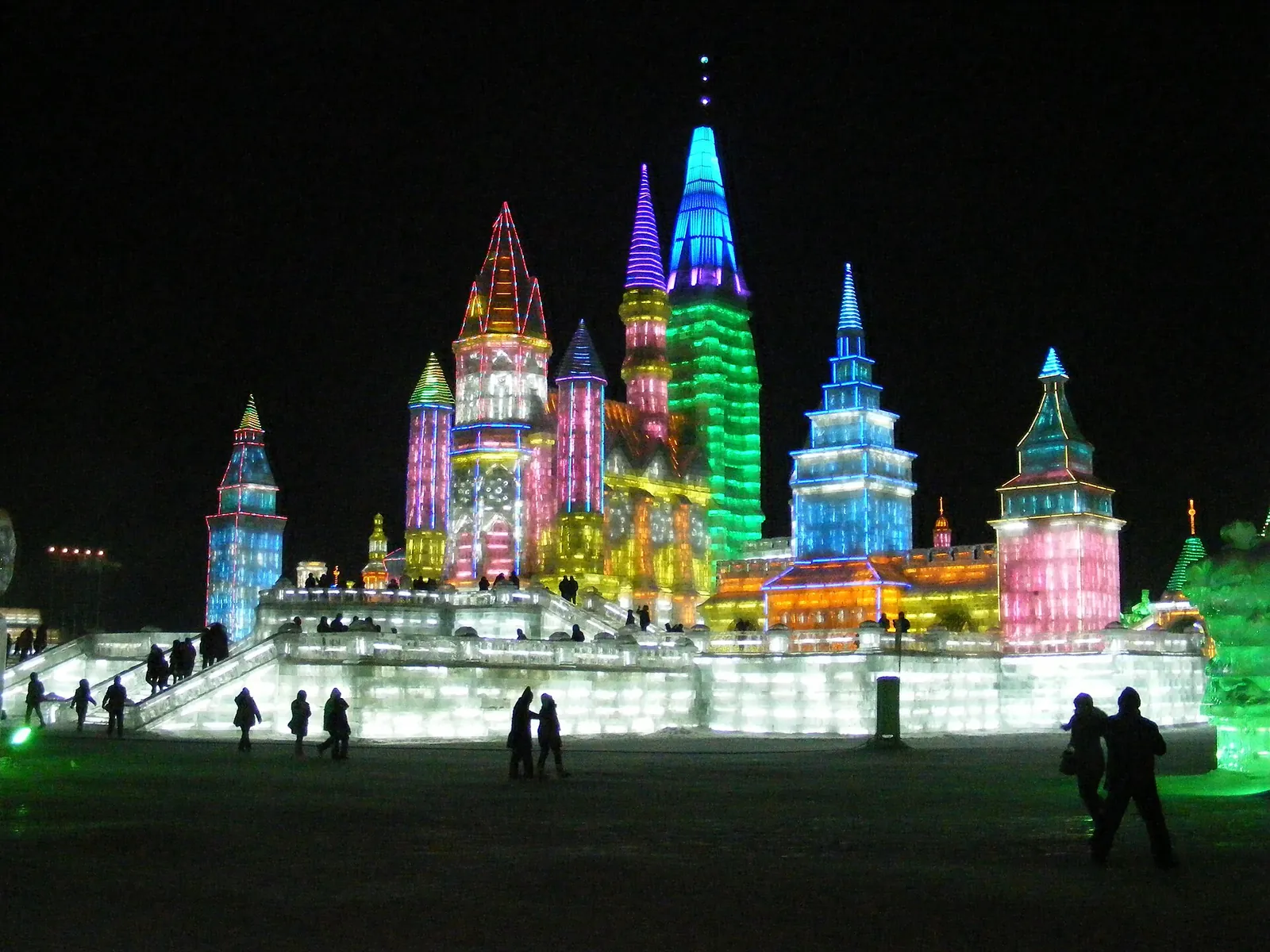 A grand ice castle at the Harbin Ice and Snow World festival, with onion-domed turrets glowing in pink, blue and purple, and visitors in heavy winter coats walking on the snow among the ice sculptures.