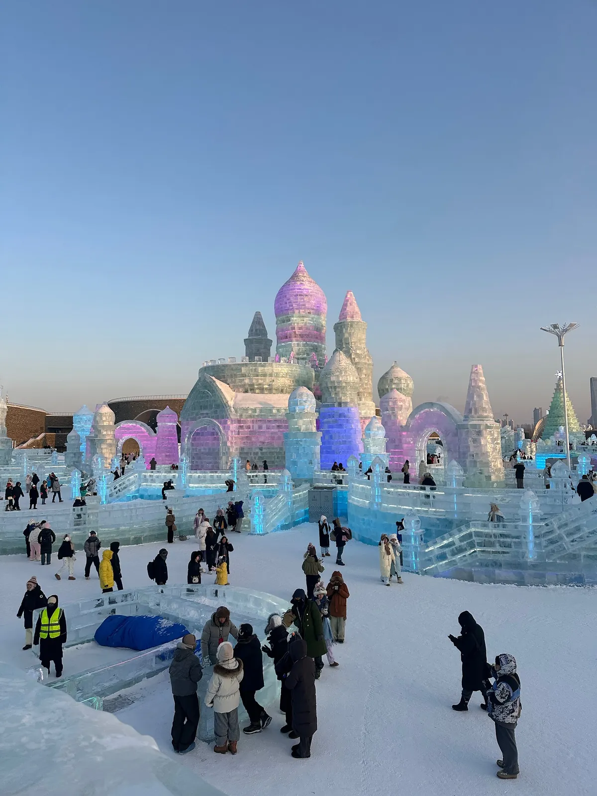 The Saint Sophia Cathedral in Harbin with a tall illuminated ice sculpture resembling a tiered crystal tower standing in front of it under a clear blue winter sky.