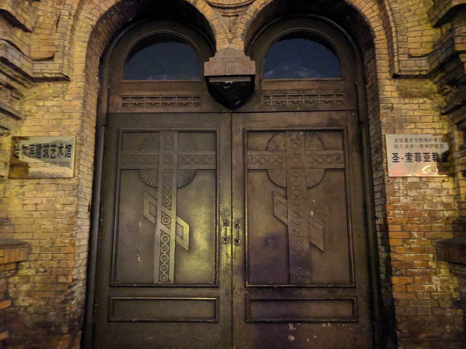 A close-up of the heavy wooden double door and brick facade of Saint Sophia Cathedral in Harbin, with cast-iron hinges and a brass plaque identifying the building as a protected cultural site.