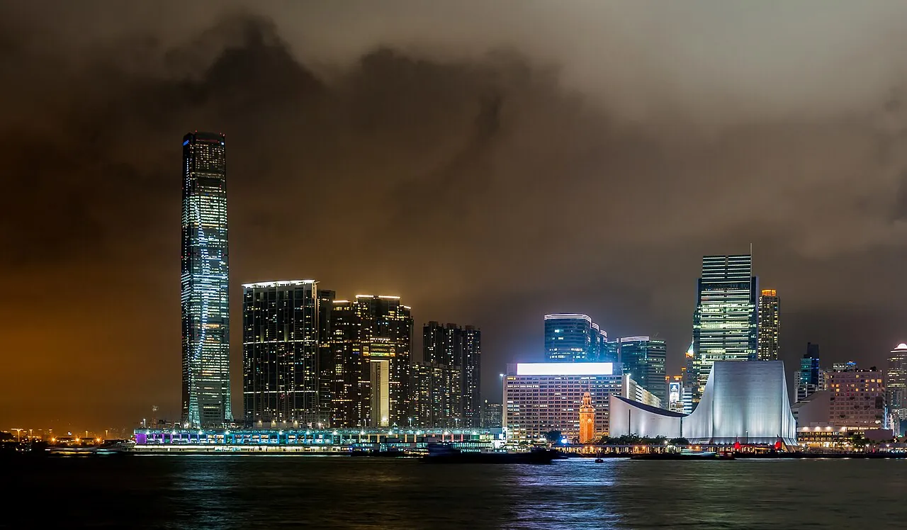 The International Commerce Centre (ICC) tower in West Kowloon lit up at night, with the Hong Kong Cultural Centre's curved roof on the right, cruise ships docked in the harbour and a dramatic cloudy sky above.