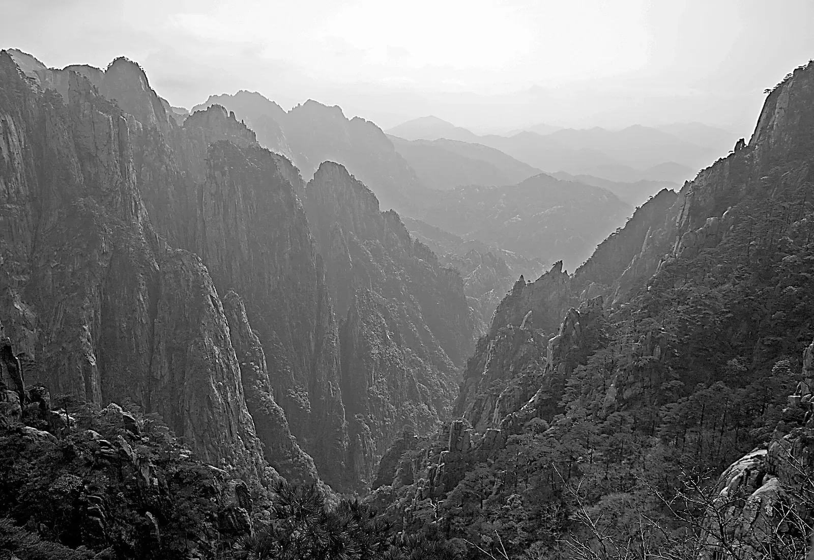 A monochrome (black-and-white) view of a misty mountain valley in Huangshan (Yellow Mountain), Anhui — with sharp granite peaks and ridges layered into the distance, a deep V-shaped valley carved into the rock, and dark pine trees covering the slopes below a hazy sky.